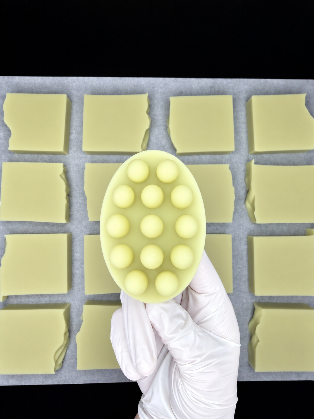 A hand wearing a glove holds a pale yellow oval green tea and tea tree oil massage soap bar with raised circular bumps, positioned over a sheet of pale yellow rectangular soap bars on a dark background.