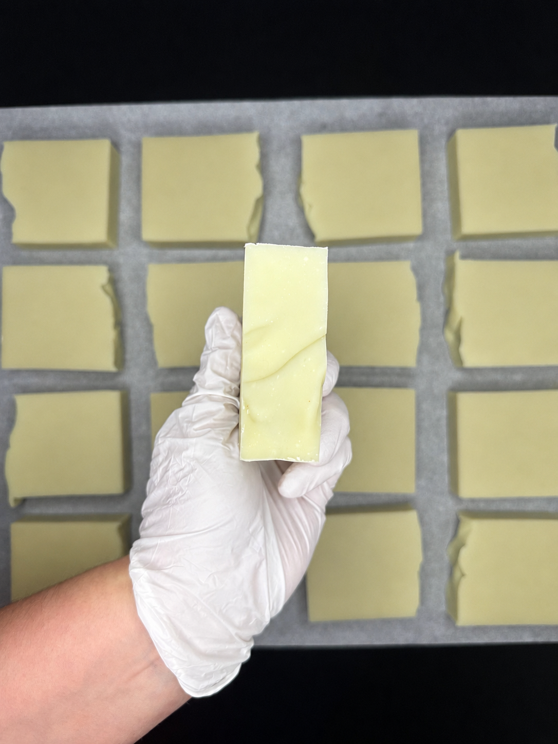 A hand wearing a white disposable glove holds a rectangular pale yellow soap bar in front of a tray of multiple matching pale yellow soap bars on a black background.