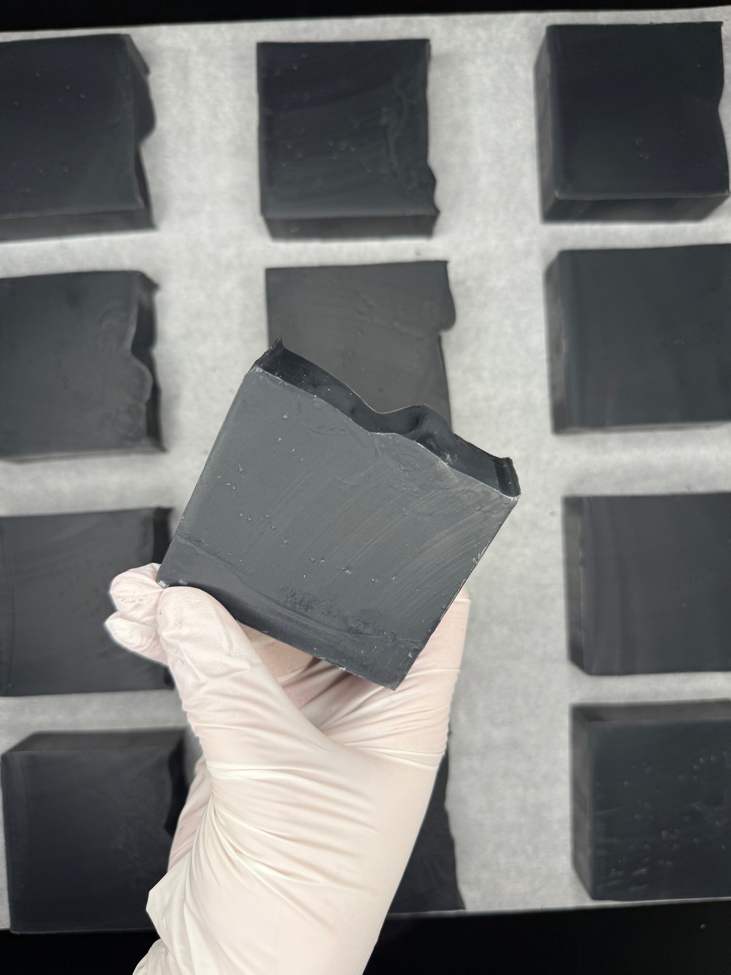 Hand wearing a glove holding a block of dark soap in front of more soap blocks.