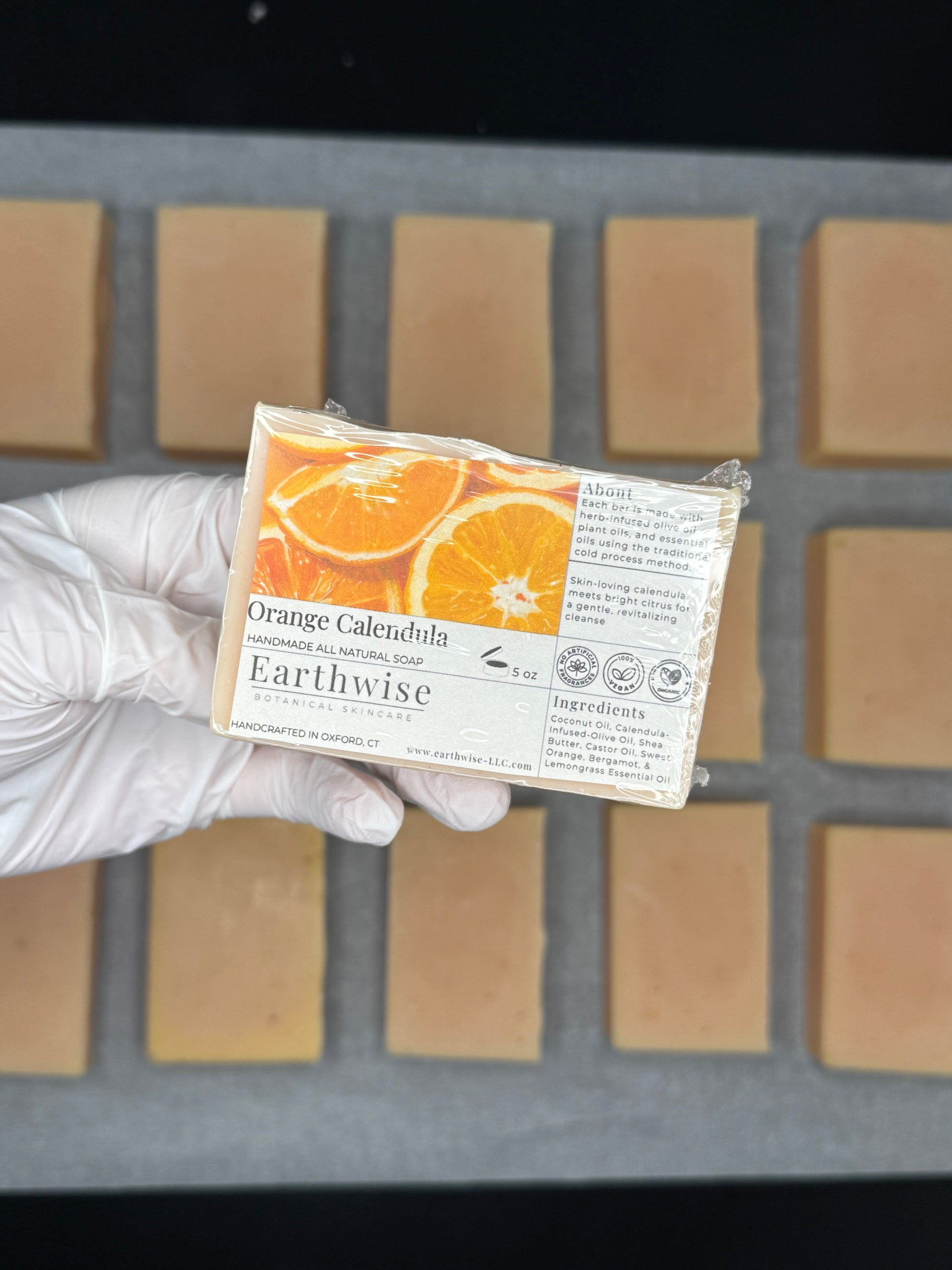 Hand holding an Earthwise Orange Calendula soap bar with a grid of soap bars in the background.