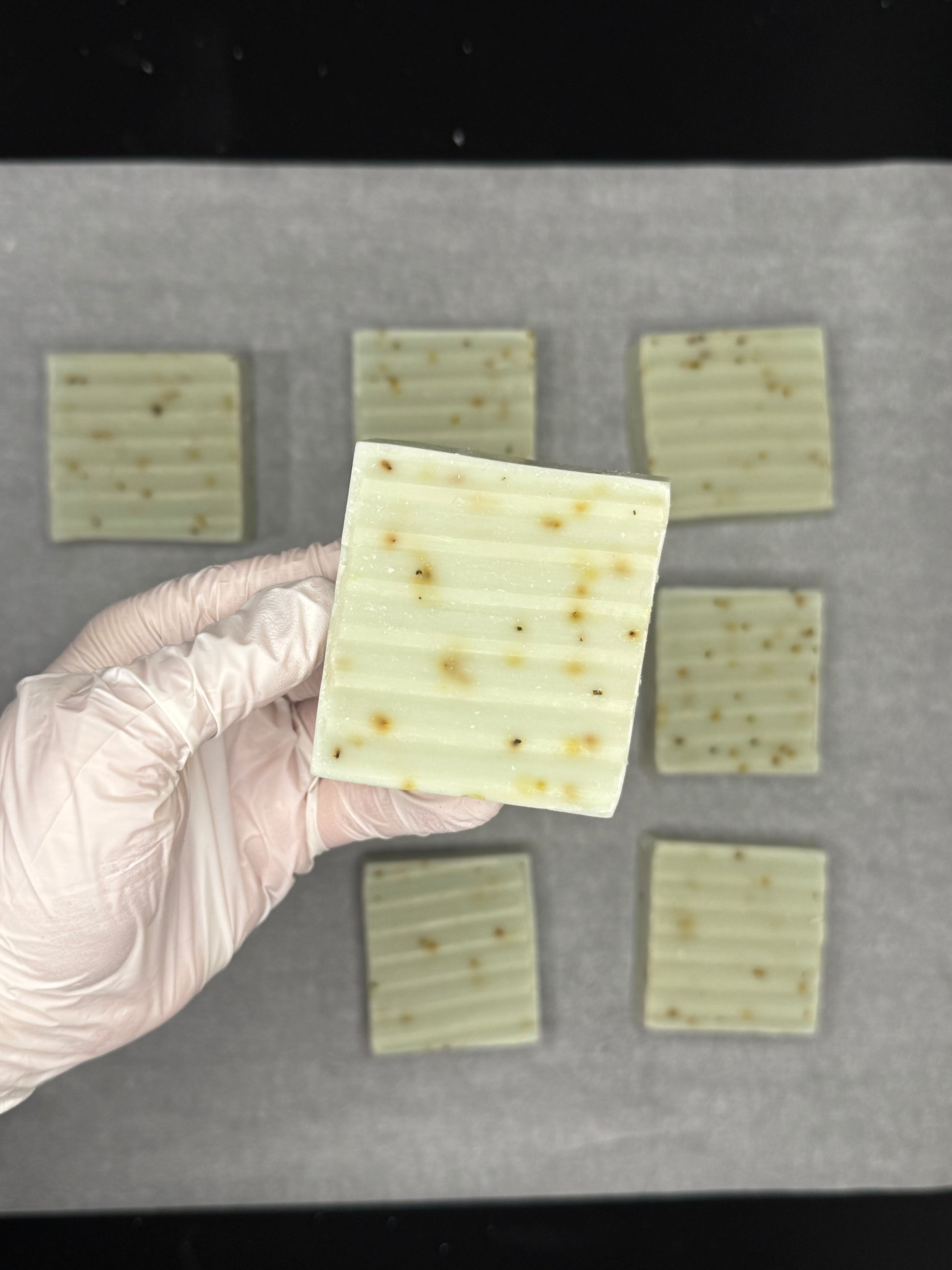 Hand wearing a glove holding a square piece of light-colored food product with visible texture, on a baking tray with more similar products.