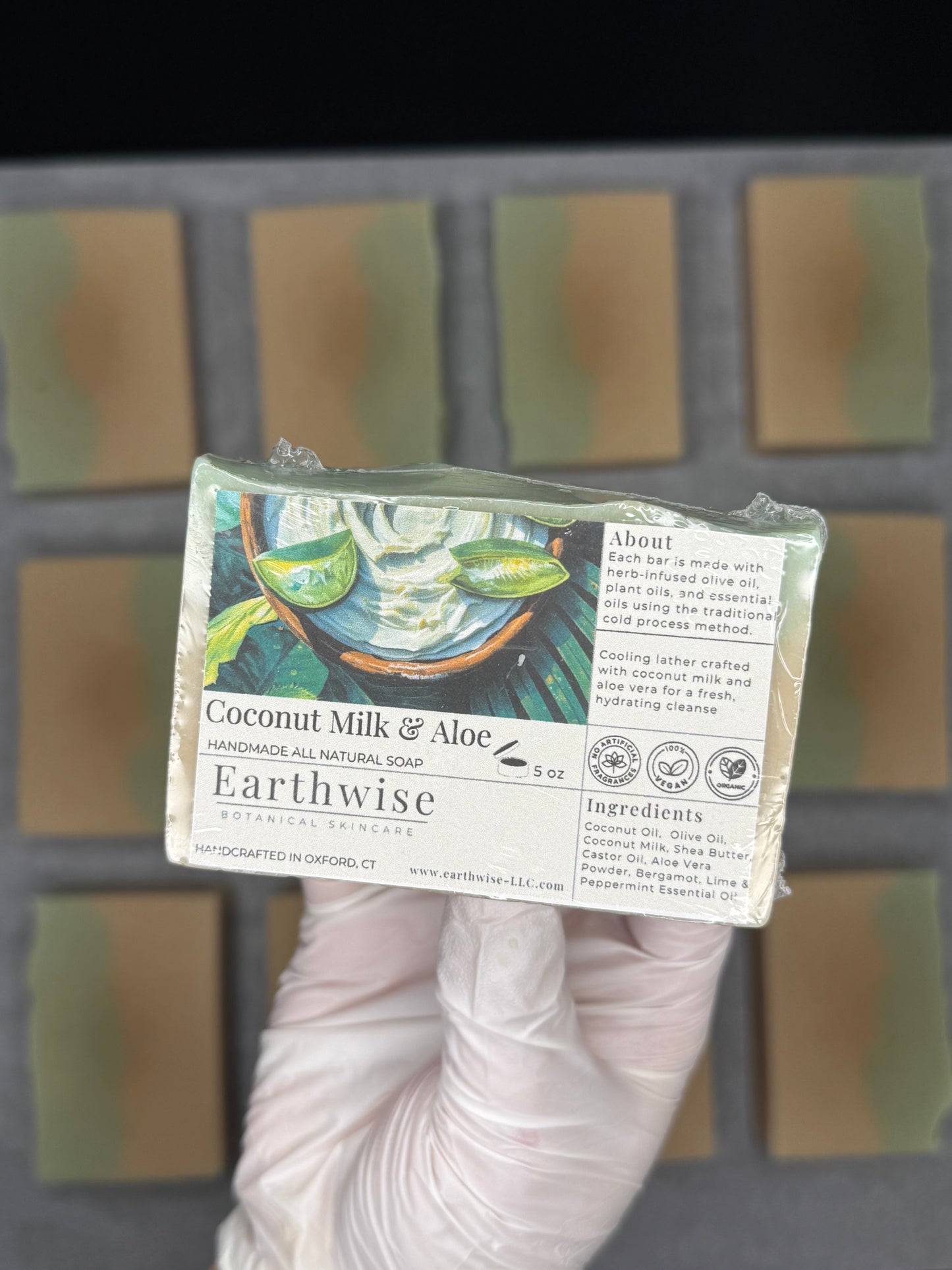Hand holding a bar of Earthwise Coconut Milk & Aloe soap with more soap bars in the background.