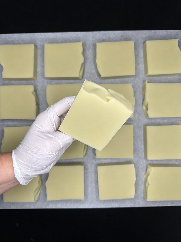 A hand wearing a white glove holds a pale yellow rectangular bar of soap over a tray lined with parchment paper, which holds multiple similar soap bars.