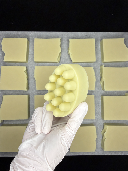 A hand wearing a white glove holds a pale yellow oval massage soap bar with raised silicone nubs, positioned in front of a tray of uncut pale yellow bars on a black background.