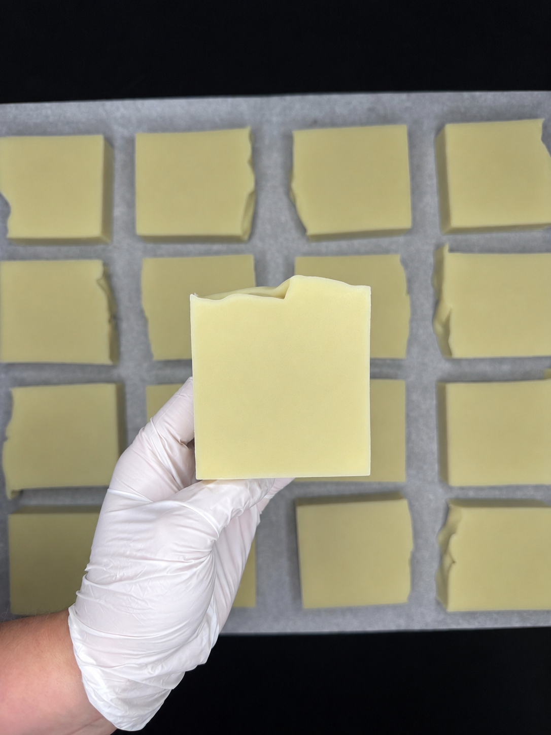 A hand wearing a white glove holds a pale yellow rectangular bar of soap in the center, with multiple similar soap bars arranged on a gray tray behind it against a black background.