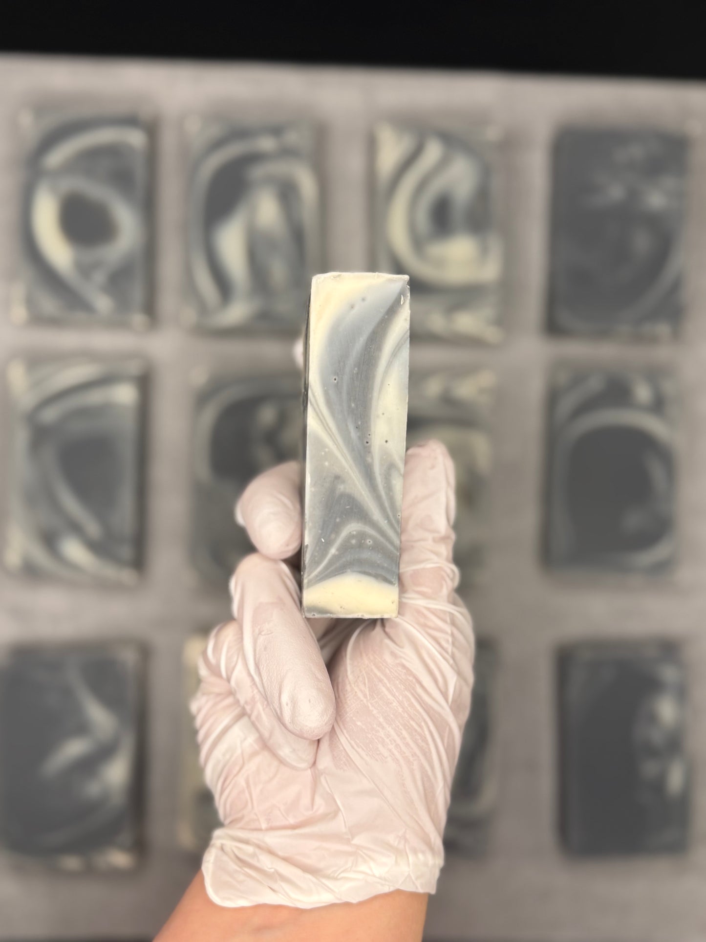 A hand wearing a white nitrile glove holds a rectangular bar of soap with a marbled gray, white, and cream swirl pattern. In the blurred background, a tray holds multiple similar soap bars.