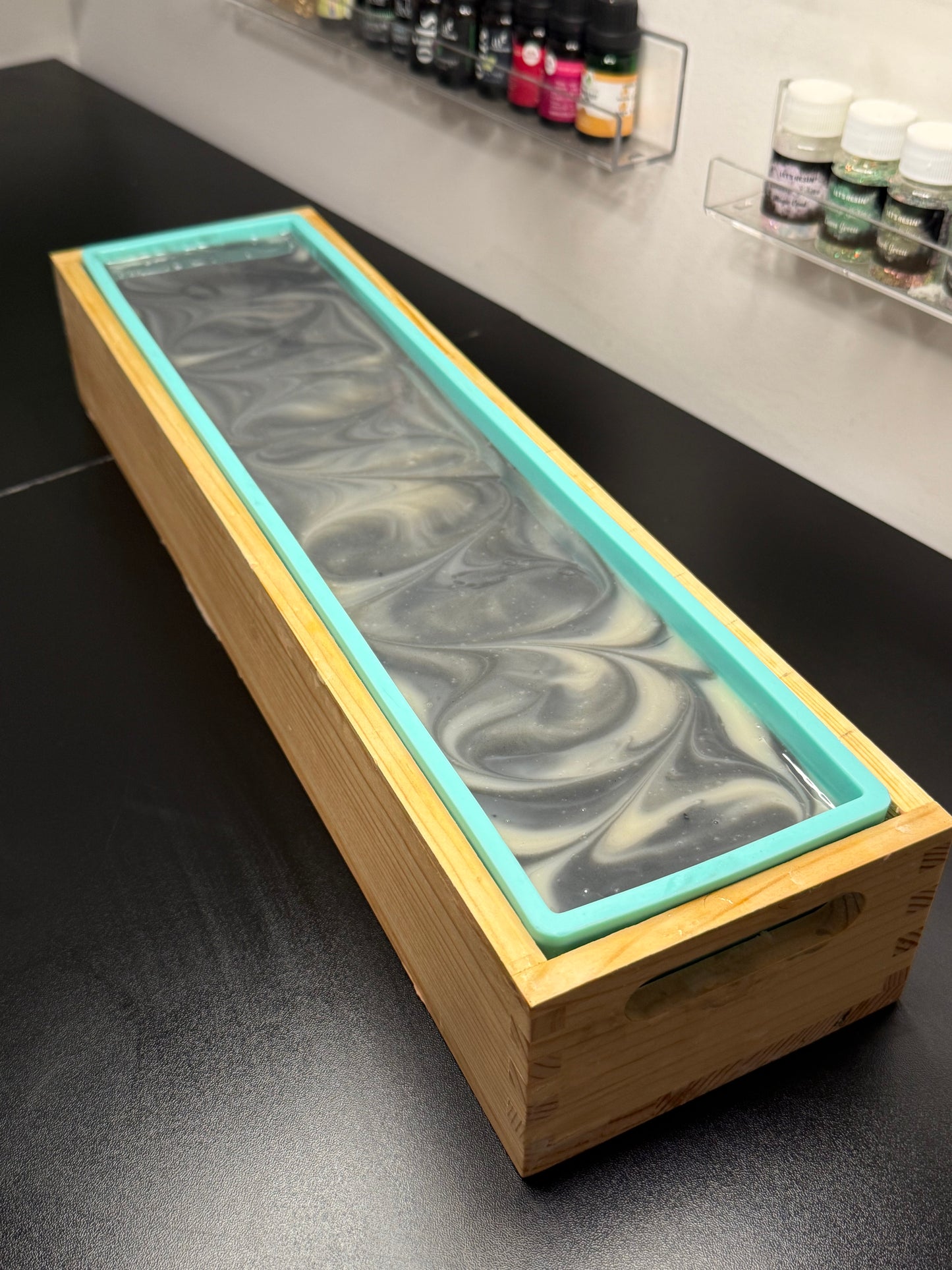 A rectangular wooden soap mold with a turquoise silicone liner and a swirled gray and white soap base, photographed on a dark tabletop with small bottles in the background.
