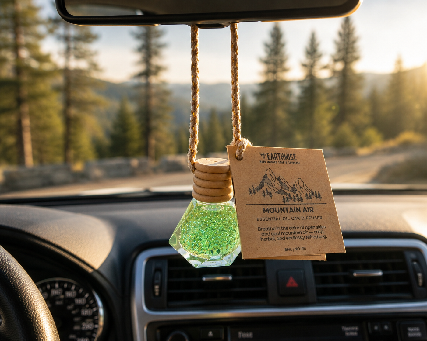 Car air freshener with mountain-themed label hanging from a car's rearview mirror with a scenic background.