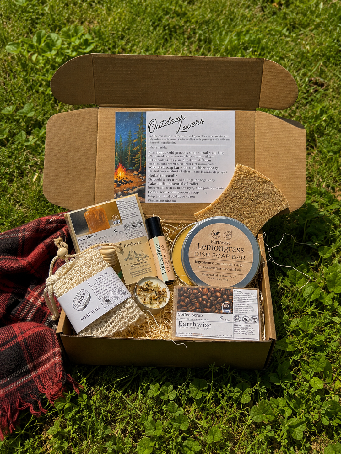 Open Earthwise Outdoor Lovers gift box displayed on green grass beside a red plaid blanket, showing handmade botanical products nestled in natural straw packing