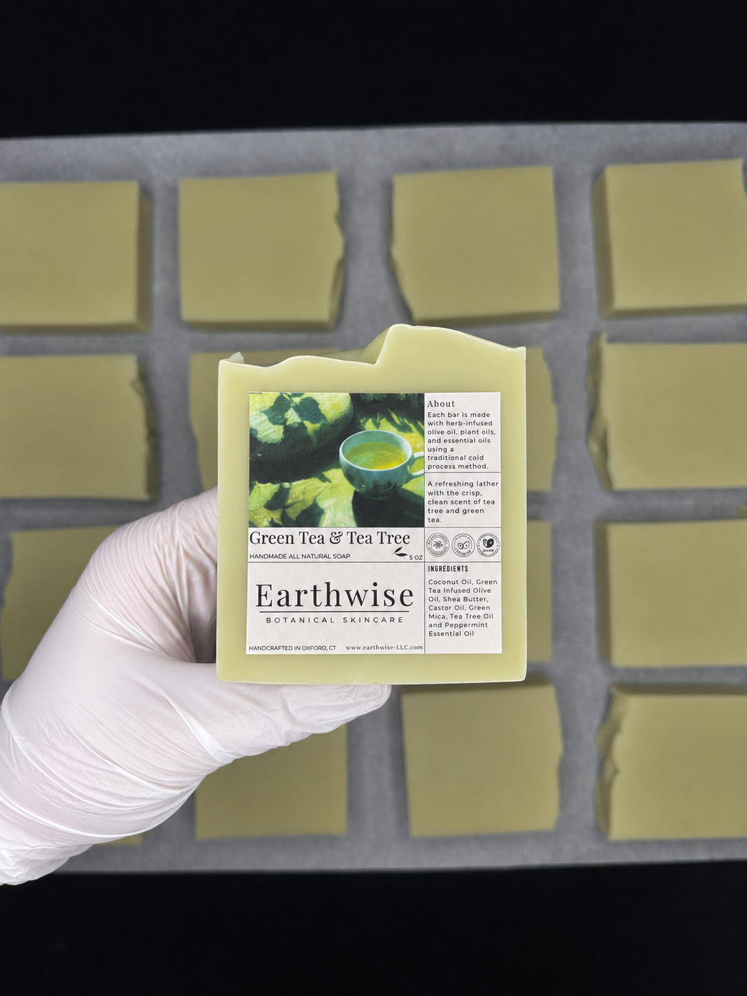 A hand wearing a white glove holds a rectangular bar of soap labeled 'Earthwise Green Tea & Tea Tree' in front of a tray of similar soap bars.