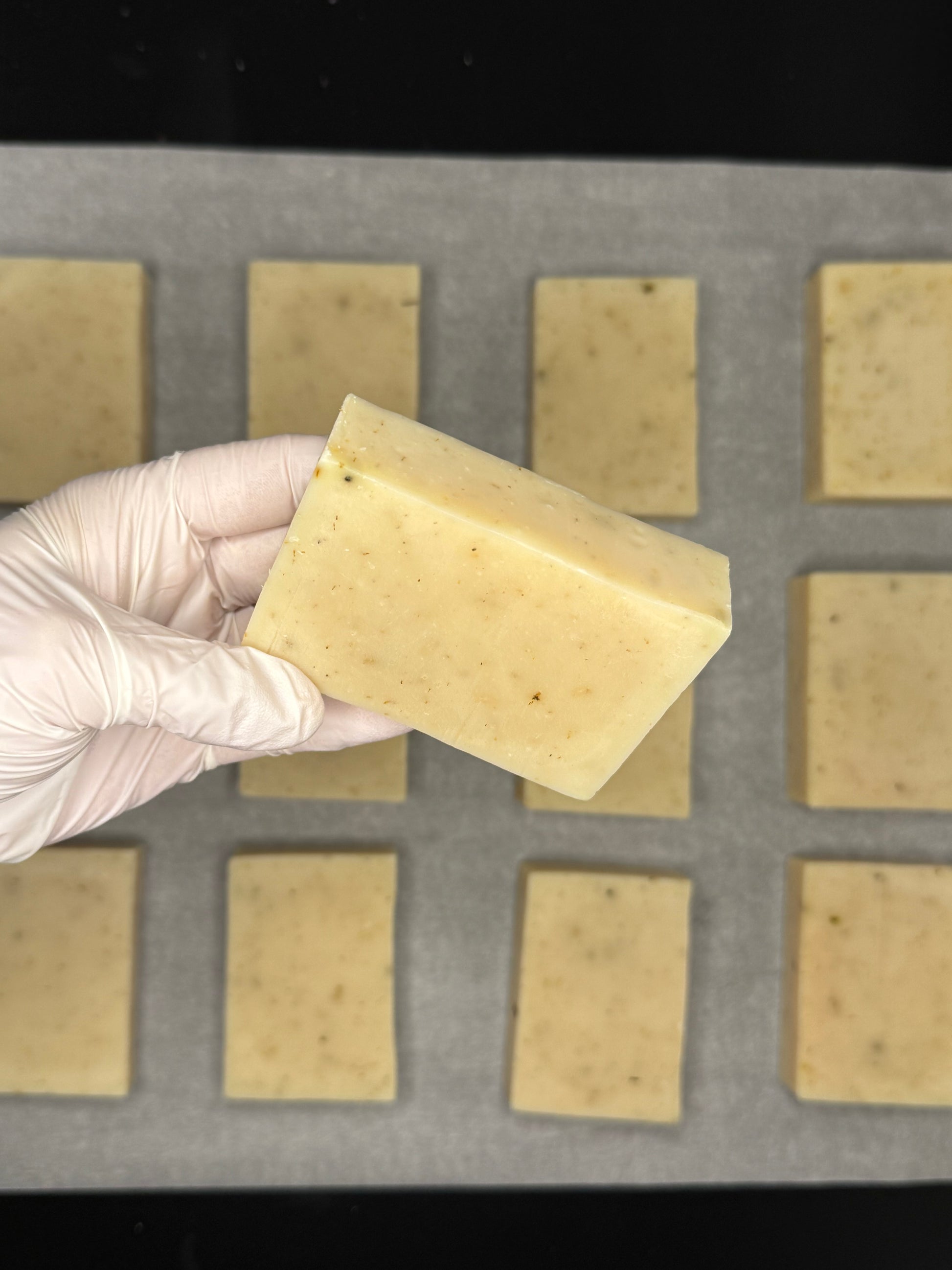 Hand wearing a glove holding a block of soap above a soap-making mold with bars already in place.