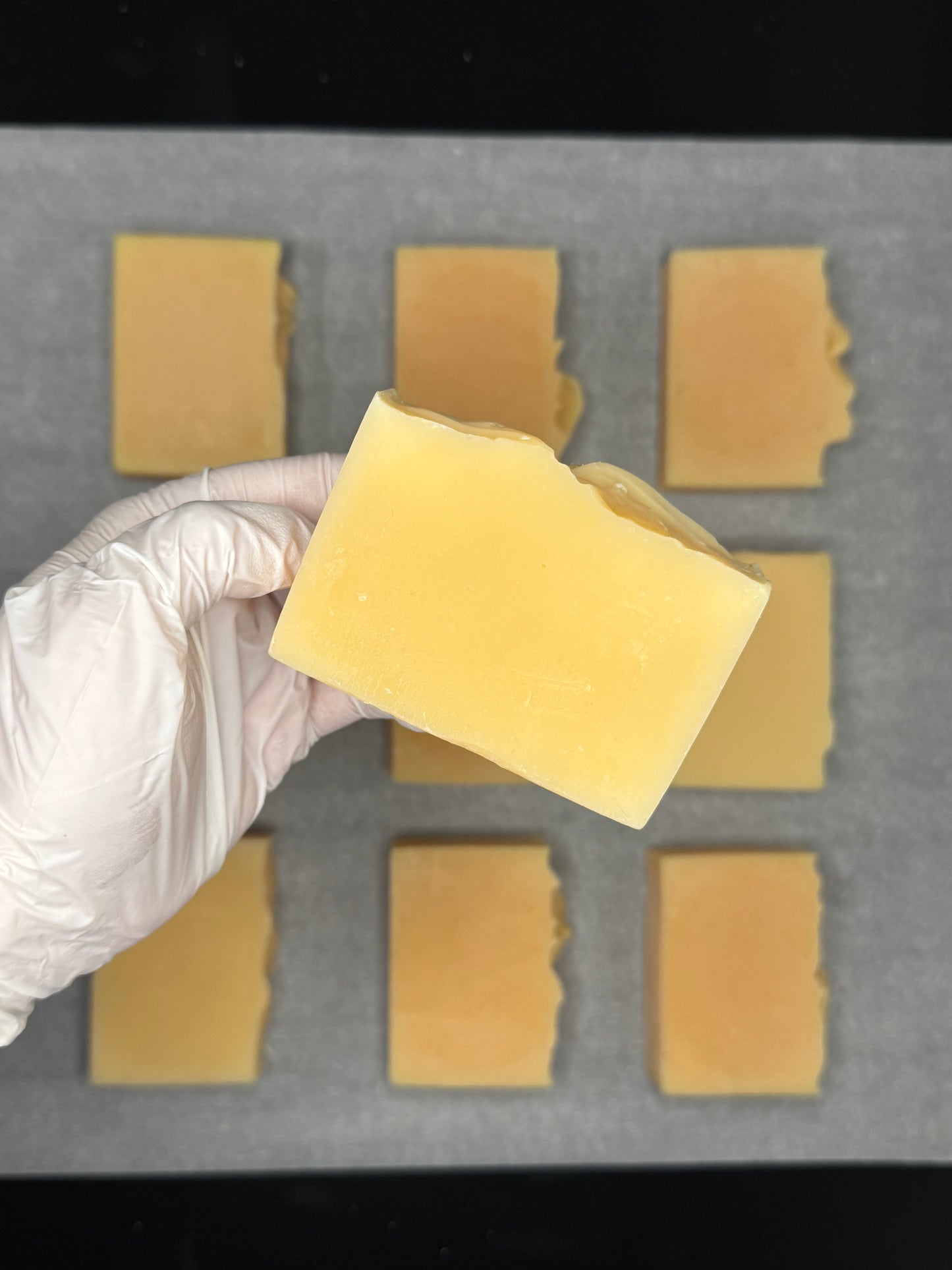 Hand wearing a white glove holding a yellow lemongrass natural soap in front of a grid of lemongrass soap bars