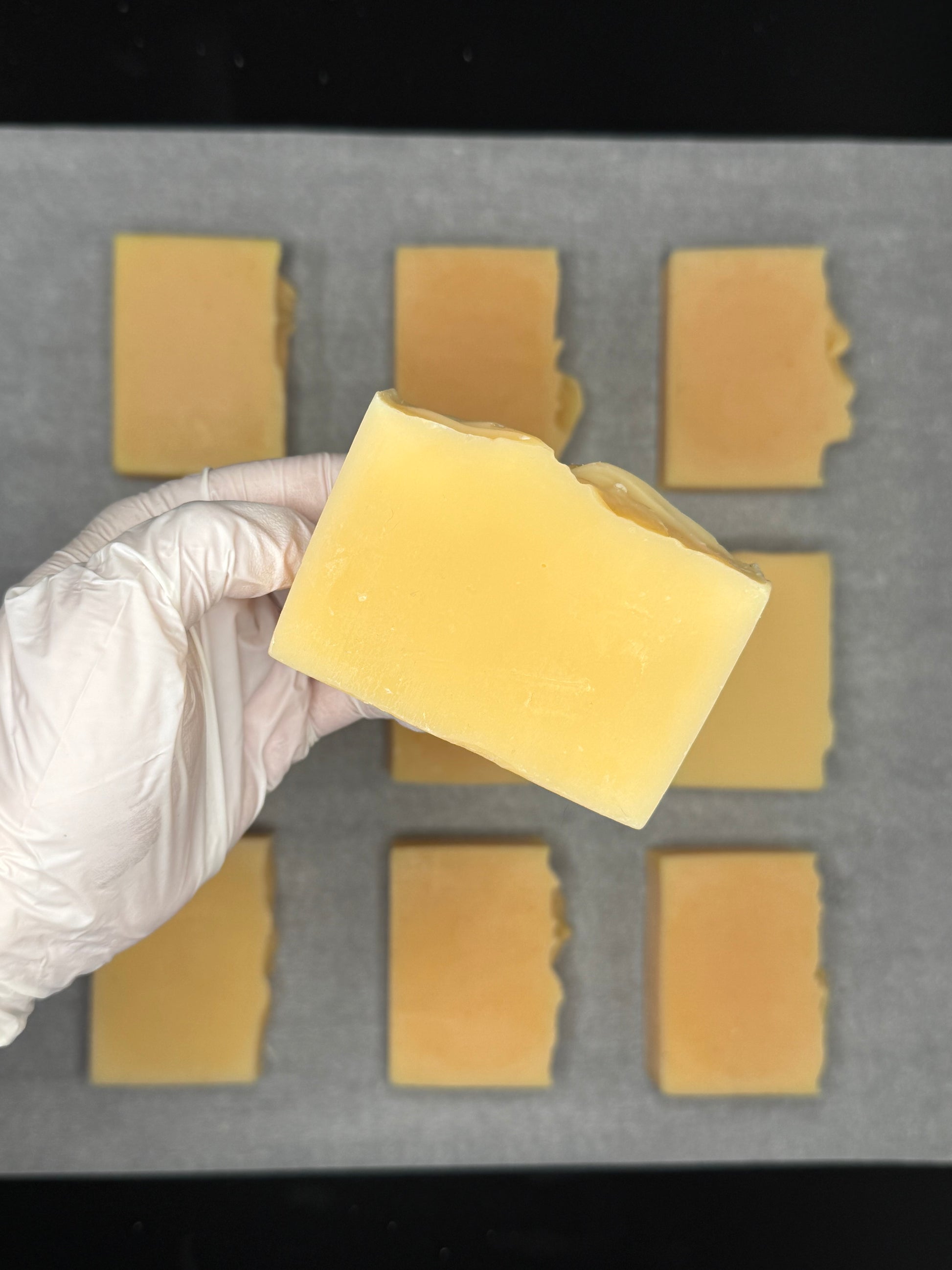 Hand wearing a white glove holding a yellow lemongrass natural soap in front of a grid of lemongrass soap bars