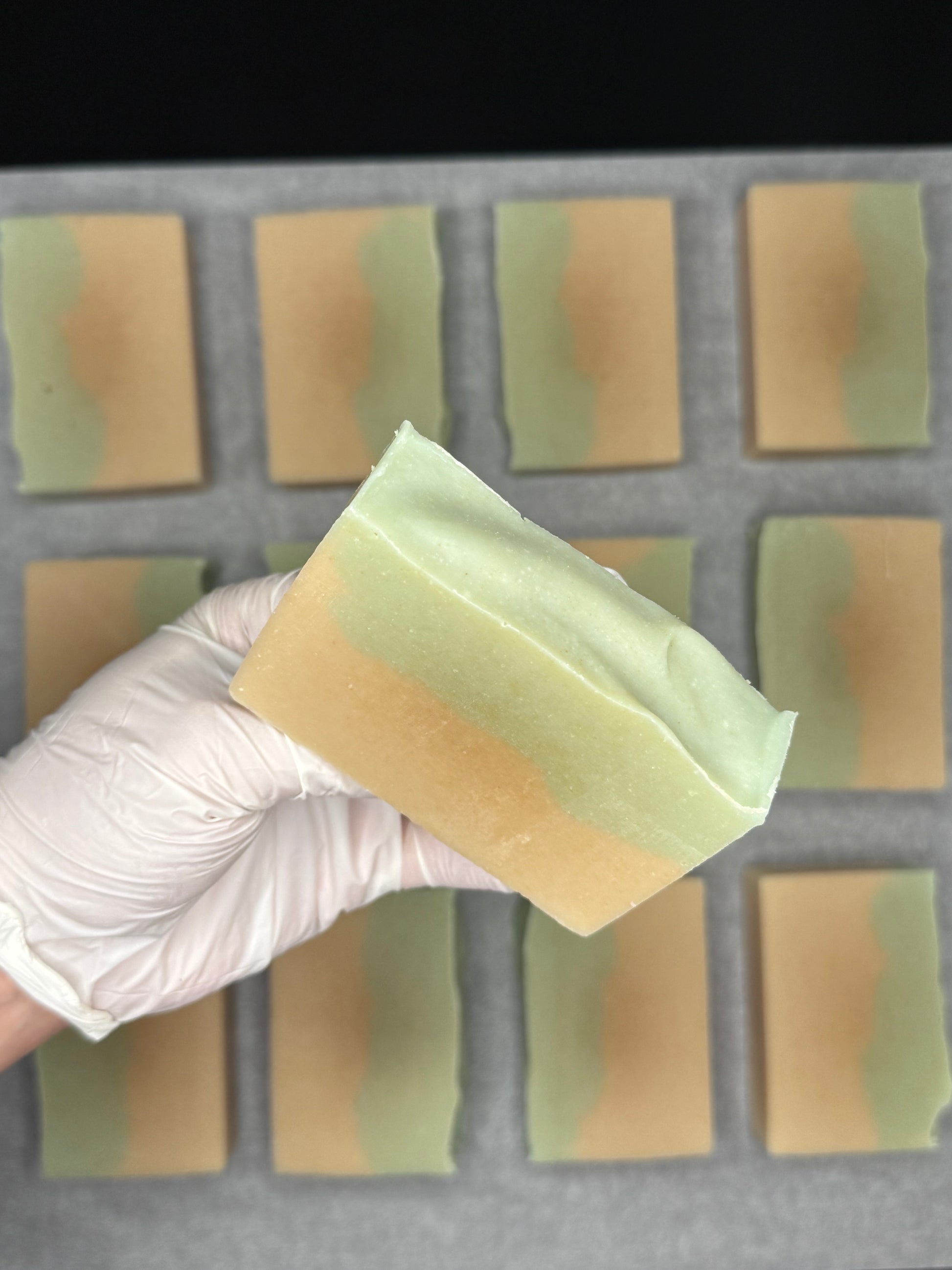 Hand holding a block of soap above a soap mold with bars in the process of being made.