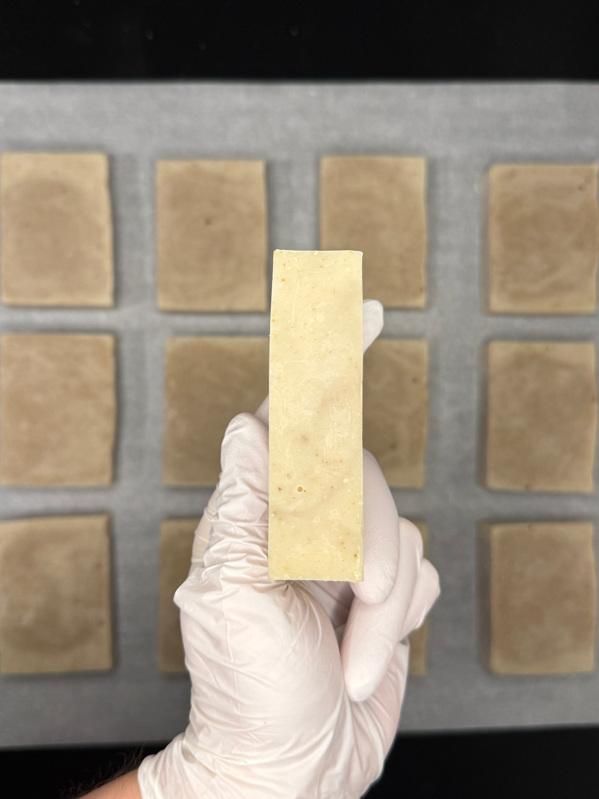 Hand holding a rectangular block of natural soap above a grid of square soap bars on a tray.