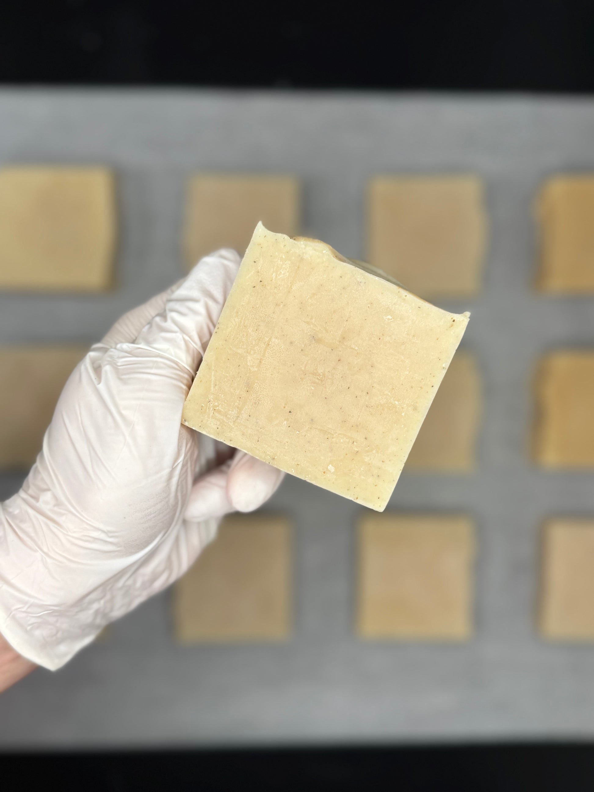 Hand wearing a white glove holding herbal face soap bar of beige material against a background with more similar pieces.