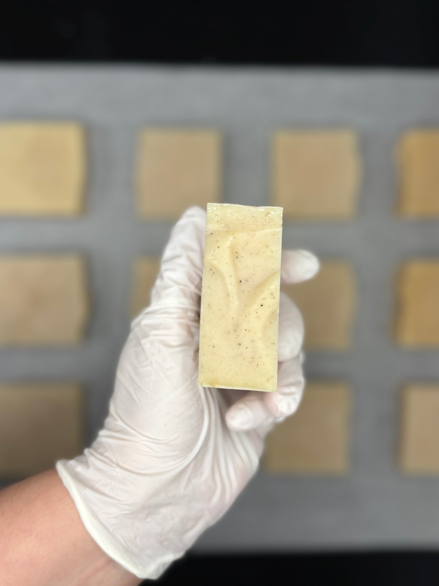 Hand wearing a white glove holding a rectangular block of soap in front of a grid of soap bars.