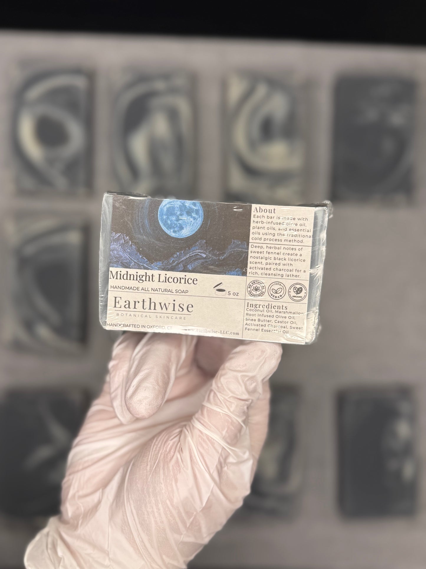 A hand wearing a translucent pink glove holds a wrapped bar of Earthwise Midnight Licorice soap against a blurred gray background of other soap bars.