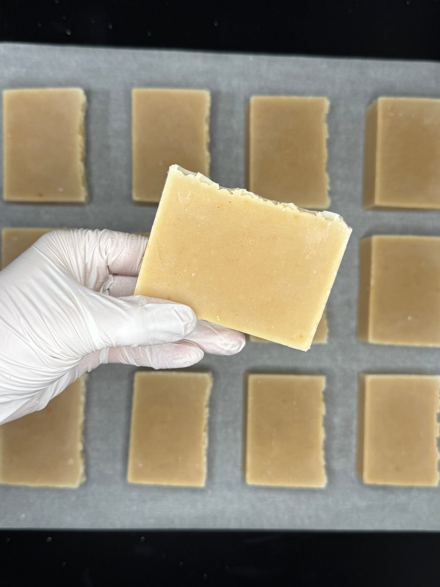 Hand wearing a white glove holding an organic honey beige soap above a soap mold with more soap bars.