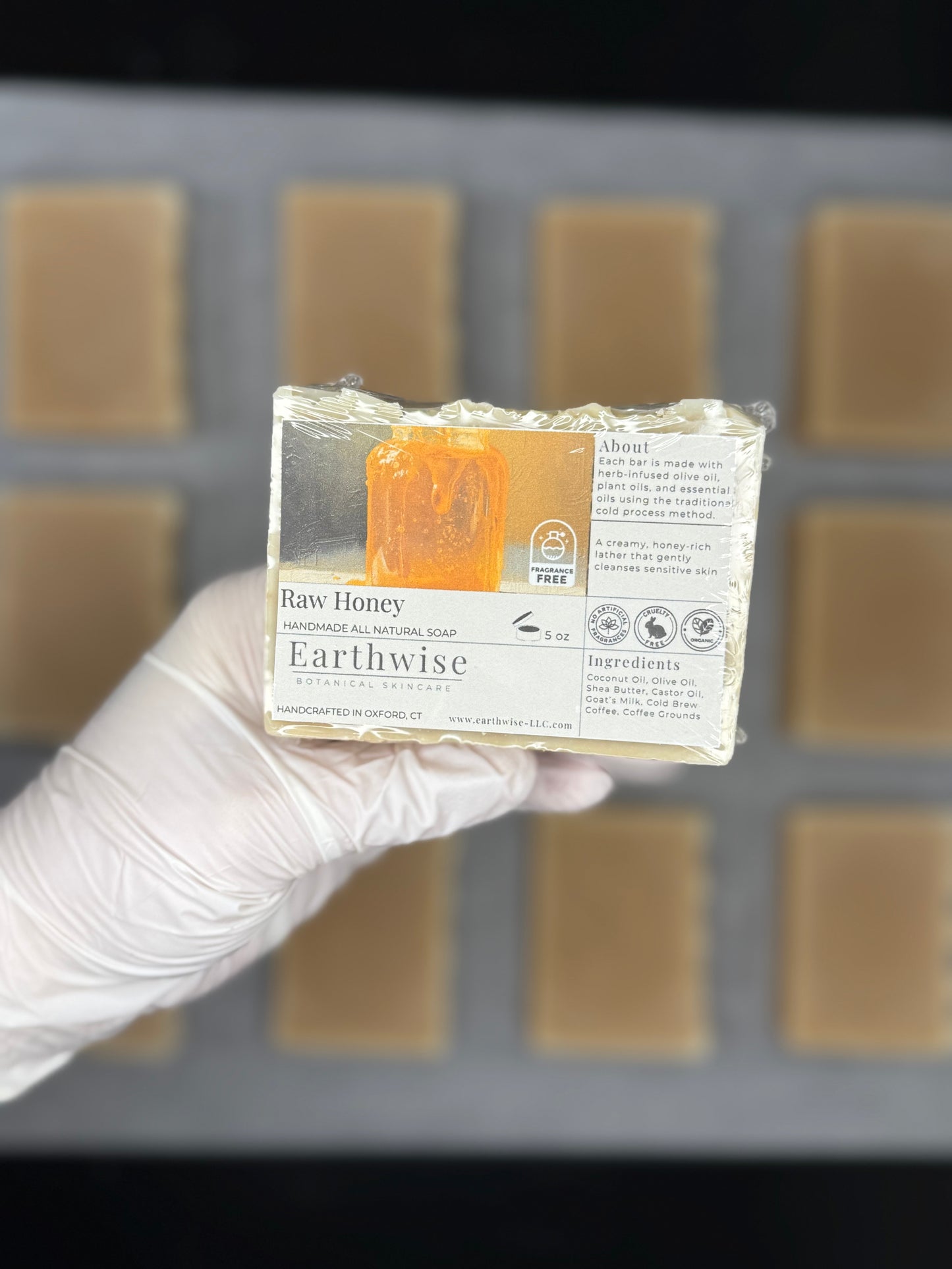 Hand holding a bar of Earthwise Raw Honey soap with a blurred soap mold in the background.