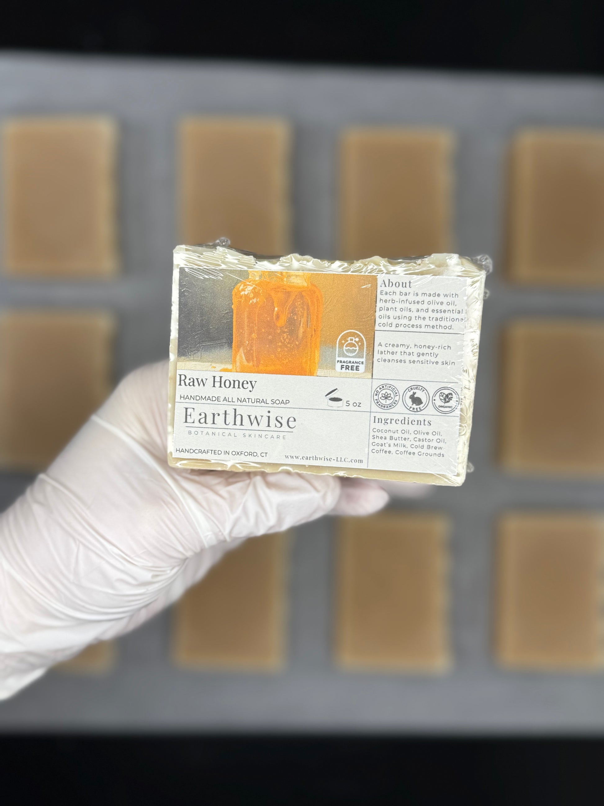 Hand holding a bar of Earthwise Raw Honey soap with a blurred soap mold in the background.