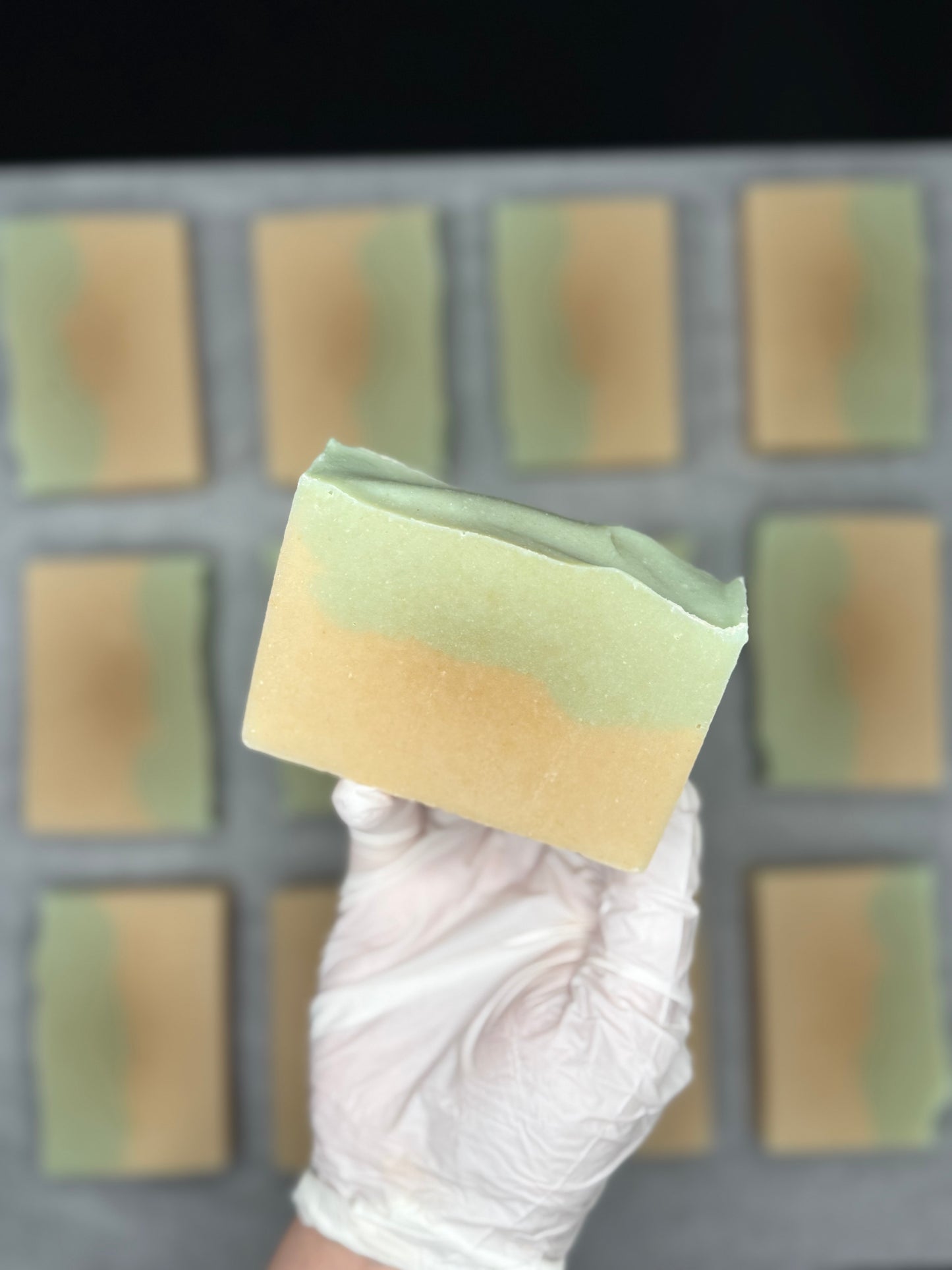 Hand wearing a white glove holding a green and yellow aloe Vera soap bar in front of a grid of more soap bars.