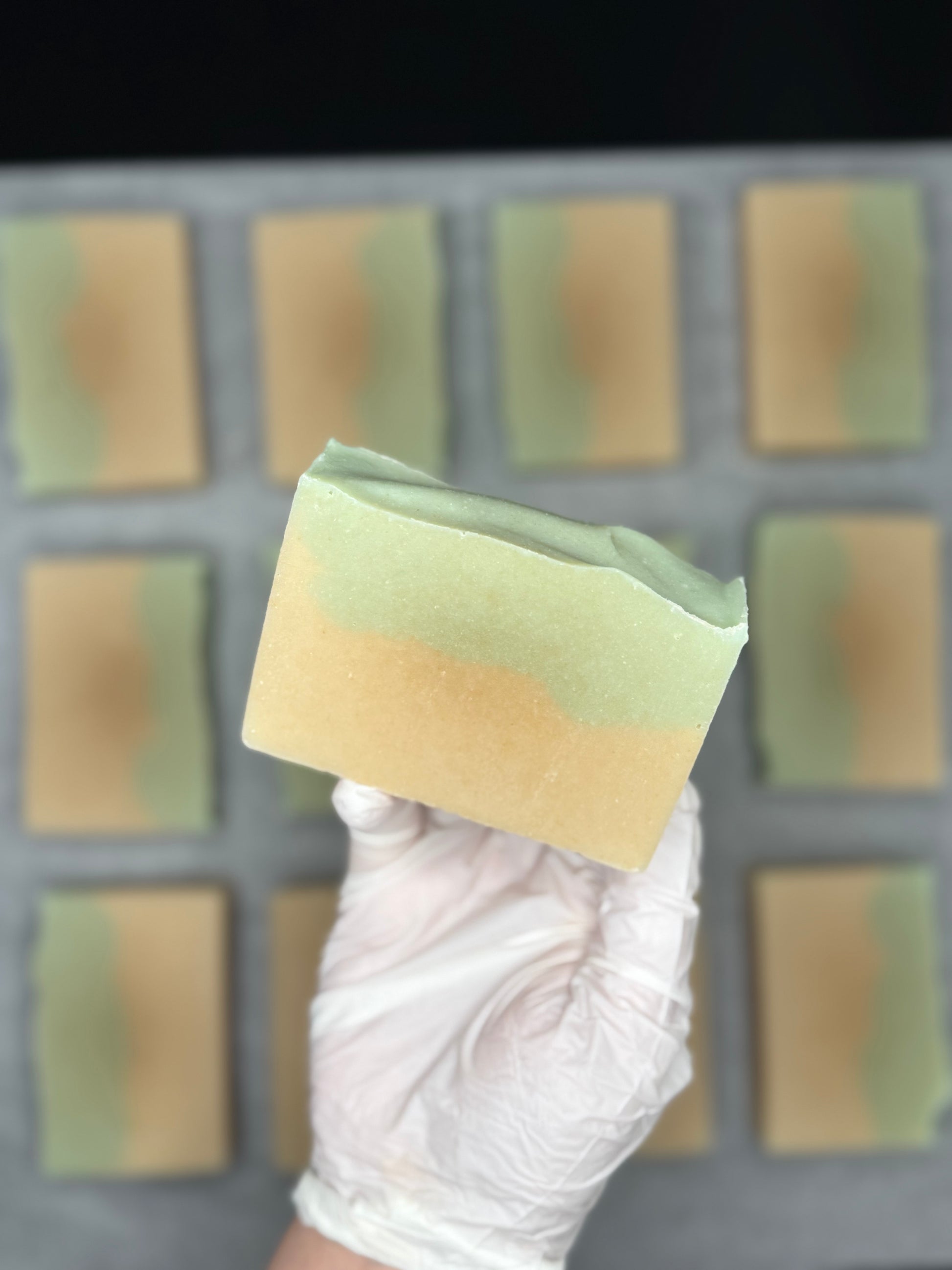 Hand wearing a white glove holding a green and yellow aloe Vera soap bar in front of a grid of more soap bars.